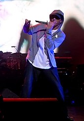 A man on a stage holding a microphone and wearing a hooded jacket, a white shirt, and blue jeans.