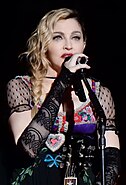 A woman with plaited hair, blue eyes and red lipstick wearing a colorful dress and guitar strap.