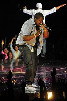 A Man wearing a white and black shirt and dark jeans, holding a microphone