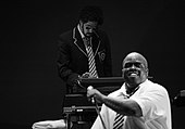 In the forefront, a man in a white dress shirt and tie holding a microphone on a stand. In the background, a man wearing a jacket, tie, and sunglasses playing keyboards.