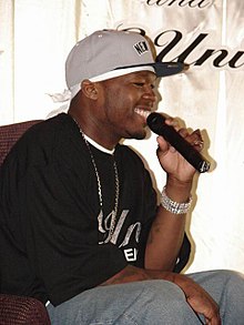 A young black man, seated, wearing a black shirt and a gray baseball cap. He is smiling and speaking into a microphone.