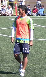 A man wearing a green T-shirt, orange bib and navy blue shorts, standing on a soccer field. In the background, people are sat watching him.