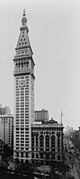 Aerial view of a thin, 50-story building with a light exterior; the building has a pyramidal roof with a large spire, and a clock is visible below the roofline.