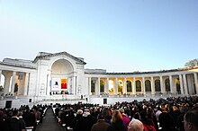 An outdoor auditorium with seated guests lined with neoclassical columns and a closed archway on one side and banners hanging inside the arch.