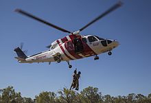 A helicopter painted red and white flying with two people wearing military uniforms hanging from a rope attached to the helicopter