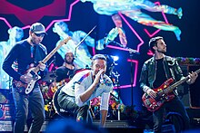 Two men play their guitars while one in the middle sings with a microphone, a fourth man is playing drums in the background