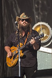 Stapleton with long brown hair wearing all-black clothing and a cowboy hat playing an acoustic guitar