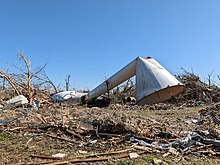 A tall white water tower lies toppled and battered on the ground, surrounded by mangled trees and strewn debris.