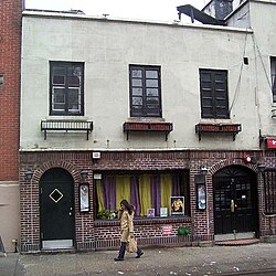 The facade of 53 Christopher Street as seen from across the street. The facade is two stories high. At the ground or first story, the facade is made of brick and consists of a wide window flanked by two archways. At the second story, there is a stucco facade with three windows, each with a flower-pot holder below.