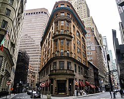 The facade of 56 Beaver Street as seen from the corner of Beaver and South William Streets. The facade is made of light orange and dark brown brick. There is a sharp curve in the center of the facade, between two streets on either side. A dark brown skyscraper is in the background.