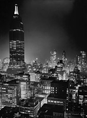 Nighttime shot of a tall skyscraper towering over smaller buildings. Most of the skyscraper is dark, with only a few floors being illuminated.