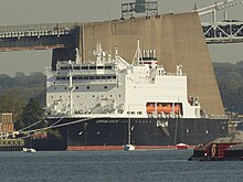 A large ship with a black hull and white superstructure. Four orange lifeboats are visible midships. Written on the bow is "EMPIRE STATE". Several small sailboats are moored in front of it, and the Throgg's Neck Bridge is visible behind.