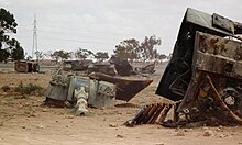 Pieces of a destroyed tank, notably the gun turret, lie on a sandy landscape.