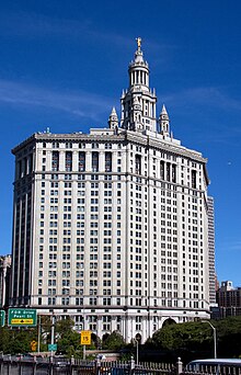 Photo of a light grey rectangular building against a piercing blue sky, on top of the building there is a tower. In the foreground of the photo is the exit sign for FDR Drive and Pearl Street. The base of the building is surrounded by large green bushes.