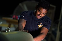 A man wearing a blue T-shirt with a military logo working on a green bomb