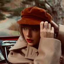 Swift in a red car, wearing a light brown trench coat and a red hat, looking back to the left.