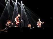 Three people performing on a stage with white lights in the background. The photo is positioned from the perspective of the crowd.