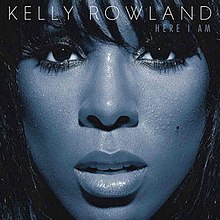 A close-up image of a woman's face covers the image. Her skin is blue and she is looking directly at the camera, with her face framed by her long black hair. Across the top in white text it reads KELLY ROWLAND (the name of the artist). On the line immediately below, in smaller text and aligned to the right, it reads HERE I AM (the name of the album).