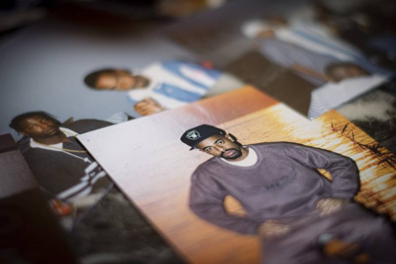 Paper photographs of young Black men lay on a table. In the top photo a man is pictured seated, wearing a dark grey sweat suit and a black Oakland Raiders cap.