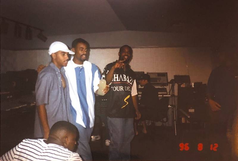 Interior of a recording studio, with 3 men standing side by side in the midfield. One man sits in the foreground with his back to the camera.