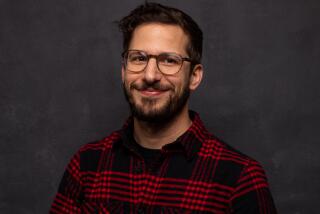 Andy Samberg smiling in glasses and a red and black plaid shirt in front of a dark background