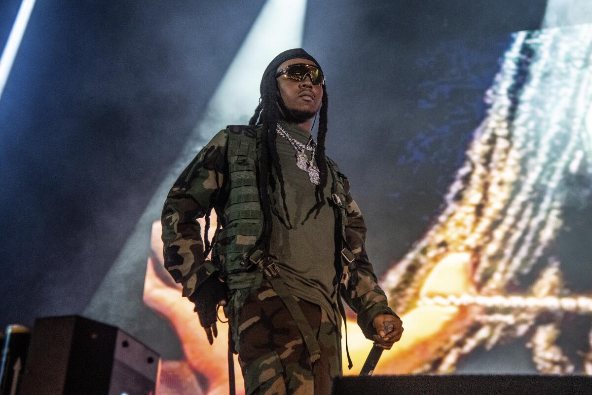 A man with long black hair wearing camouflage and holding a microphone on a stage.