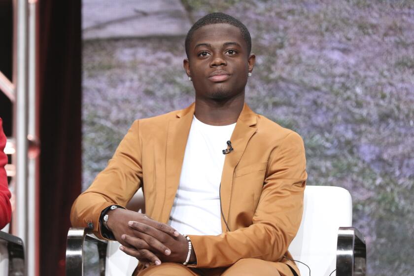 A Black man in a tan suit and a white t-shirt sitting in a white chair with his hands on the armrest