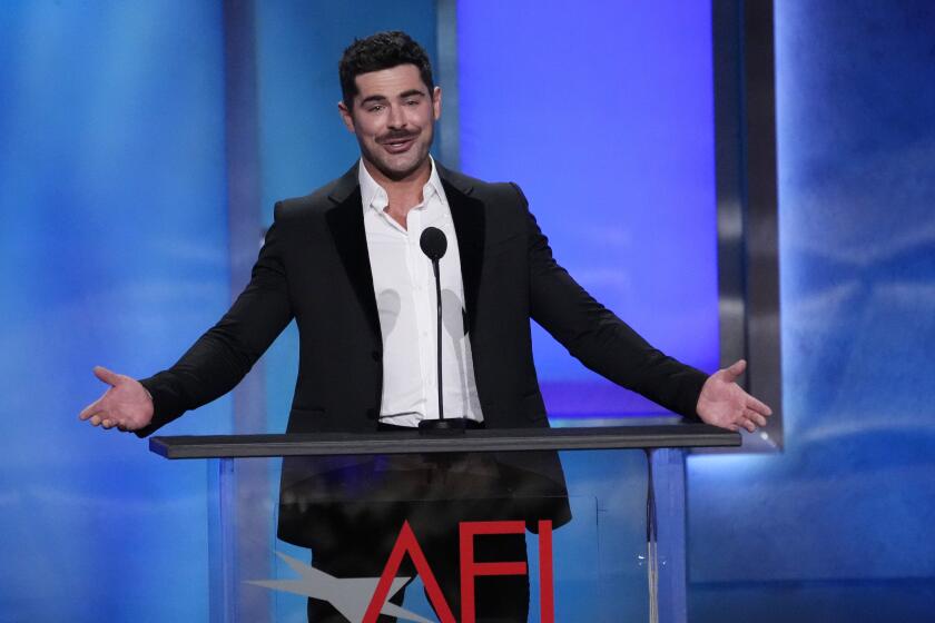 Actor Zac Efron standing with his hands streched from his sides, wearing a white button up shirt and dark blazer. 