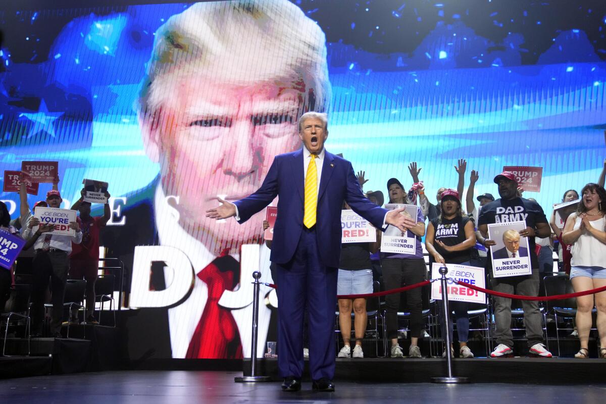 Donald Trump speaks, a huge image of himself as a backdrop, as supporters hold signs, one reading "Joe Biden, you're fired"