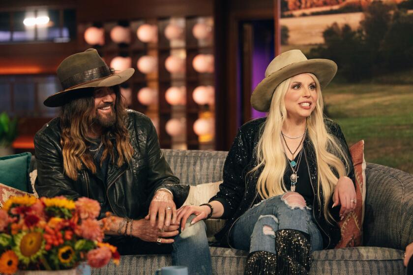 Billy Ray Cyrus with long hair wearing a wide-brimmed hat and jeans sitting next to a blonde woman in a similar outfit