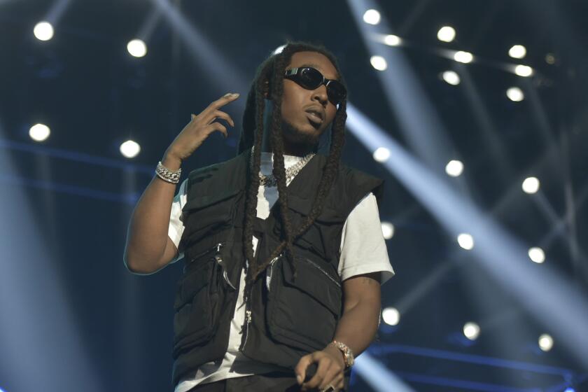 A man with long black hair wearing a black vest and white T-shirt on a stage