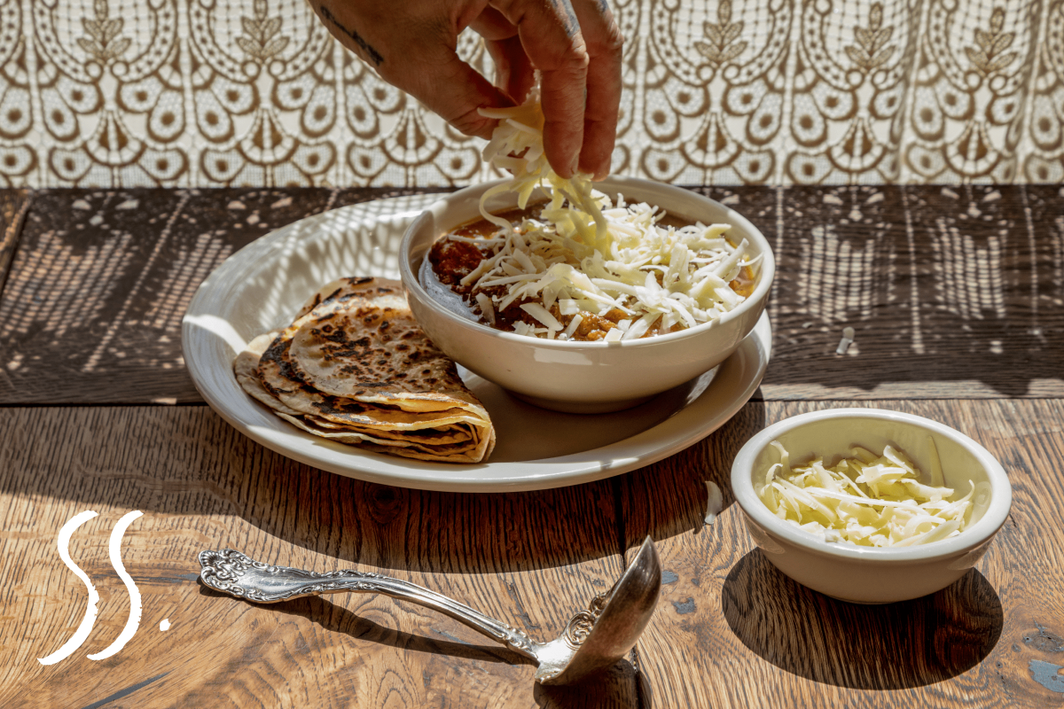 #55: A hand putting graded cheese on top of Green Chili Stew and Tortillas