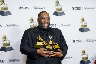 Killer Mike in a black suit holding three golden Grammy Awards in his arms as he smiles and poses for pictures