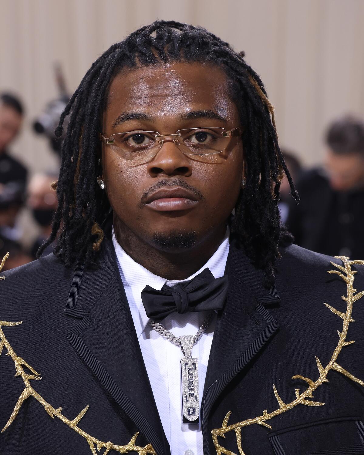 A man with short braids in a dark suit with an embroidered motif that looks like a thorny branch. 