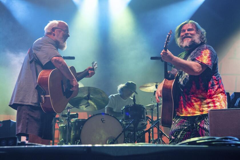 Kyle Gass and Jack Black both playing guitars on opposite ends of a stage in front of a man on a drumkit