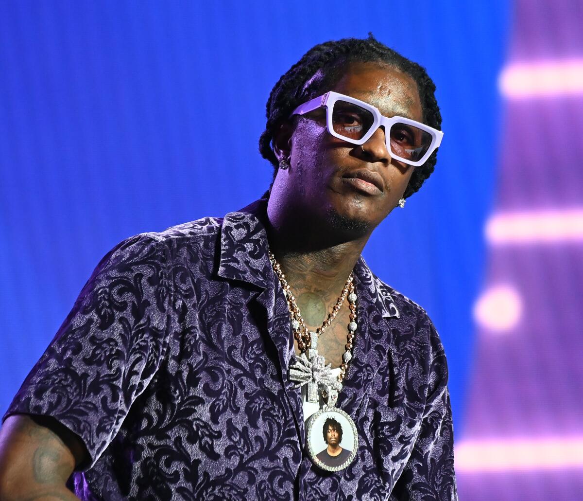 Young Thug leans forward and looks ahead while clad in thick white glasses, a dark patterned shirt and multiple necklaces