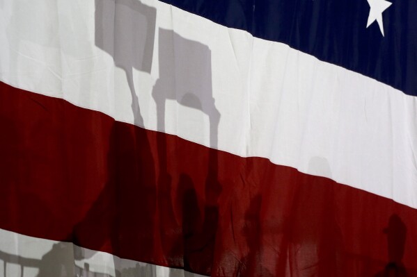 Supporters of Democratic presidential candidate Hillary Clinton cast shadows on an American flag behind them while listening to speakers while waiting for Clinton's arrival at a rally, Wednesday, March 2, 2016, in New York. (AP Photo/Julie Jacobson)