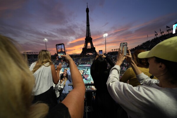 Spectators photograph a colorful sunset at Eiffel Tower Stadium prior to a beach volleyball match between The United States and Canada at the 2024 Summer Olympics, Saturday, July 27, 2024, in Paris, France. (AP Photo/Robert F. Bukaty)