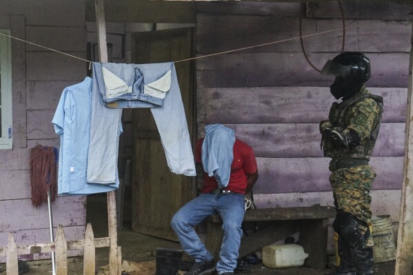 A detainee sits with their hands cuffed by a member of the National Border Service (SENAFRONT) on the outskirts of Santa Fe in the Darien province of Panama, Wednesday, Aug. 7, 2024. Panama's border police arrested alleged human traffickers of migrants who enter through the Darien jungle on their way to the U.S. (AP Photo/Abraham Teran)