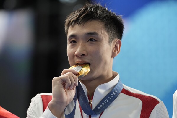 Gold medalist China's Cao Yuan bites his gold medal on the podium after the men's 10m platform diving final, at the 2024 Summer Olympics, Saturday, Aug. 10, 2024, in Saint-Denis, France. (AP Photo/Lee Jin-man)