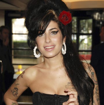 amy winehouse smiles at the camera, she wears a black strapless top with large white hoop earrings and a red rose in her beehive hairdo