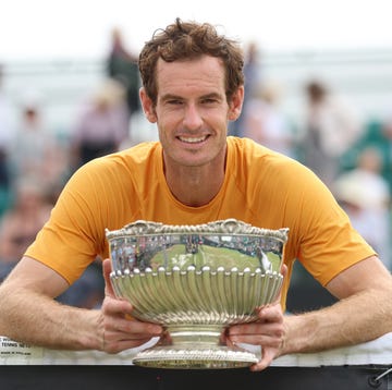 andy murray smiles at the camera while holding a silver bowl trophy, he wears an orange t shirt and leans against a tennis net