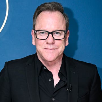 kiefer sutherland smiles at the camera, he wears black glasses, a black suit jacket and a black collared button up shirt