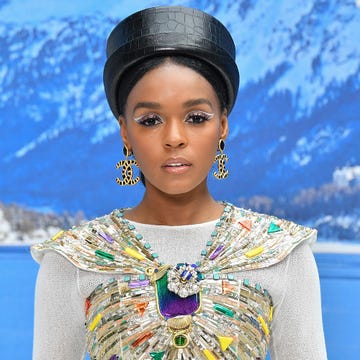 janelle monae looks at the camera, she wears a bejeweled top with a white mesh long sleeve shirt underneath, a black top hat, and large gold and black dangling earrings