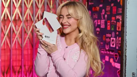 Official Charts Company Sabrina Carpenter holding up her number one trophy from the Official Charts Company
