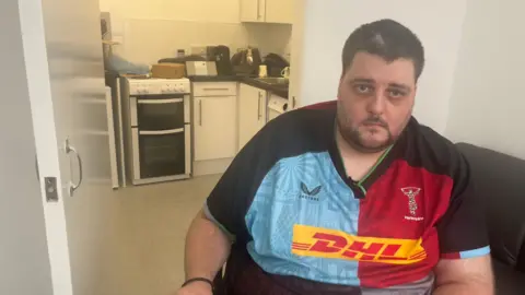 Nathan Rollinson, a man in a red and blue rugby shirt, sitting in front of his kitchen with a cooker that opens onto the floor