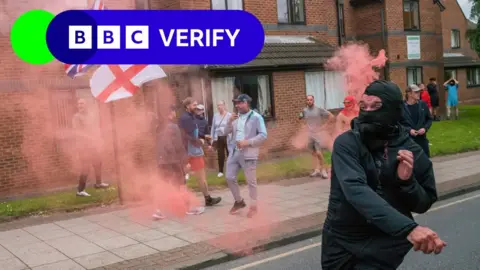 Police officers wearing riot gear in Sunderland, viewed from behind close to the camera, as in the middle distance an overturned car burns and a crowd mills around