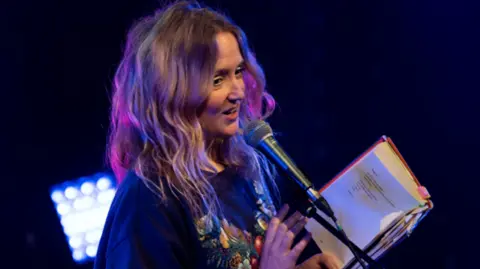 Poet Hollie McNish on stage, she is talking into a microphone while holding a book, she is wearing a blue top with a pattern/graphic on the front