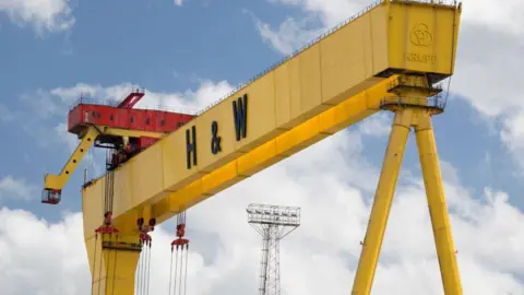 A close shot of the top of one of the large yellow Harland and Wolff cranes in Belfast. The red operator box is on the left hand side of the image. The sky in the background is light blue with white clouds.