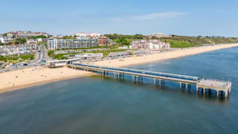 A pier extends out over the sea, beyond a sandy beach. There is a smattering of people on the beach.